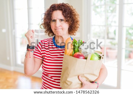 Senior woman holding paper bag full of fresh groceries from the supermarket annoyed and frustrated shouting with anger, crazy and yelling with raised hand, anger concept