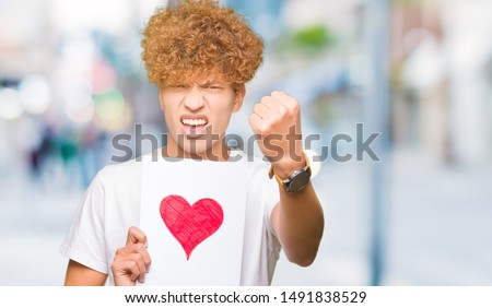 Young handsome man holding paper with red heart annoyed and frustrated shouting with anger, crazy and yelling with raised hand, anger concept