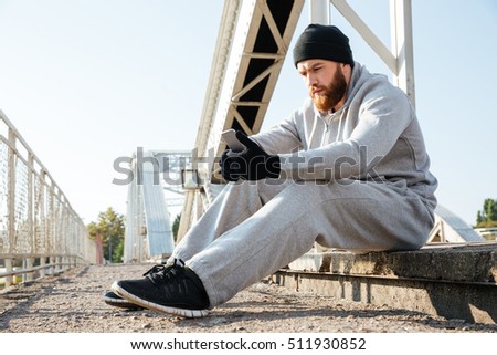 Portrait of a young sports man in hat and sports wear using mobile phone while having rest after workout outdoors
