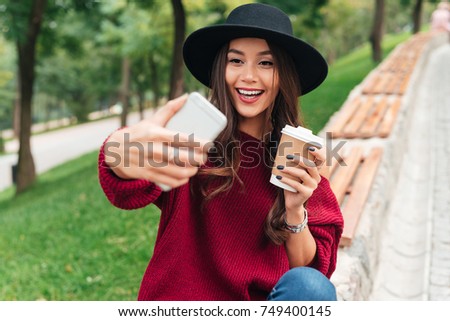 Retrato de una feliz joven asiática vestida con sombrero y suéter sosteniendo una taza de café mientras se sienta en un banco y tomando un selfie al aire libre