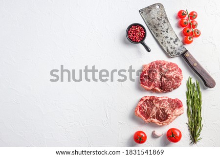 Raw top blade steak, organic meat with seasonings, rosemary and butcher cleaver. White textured background. Top view with space for text.