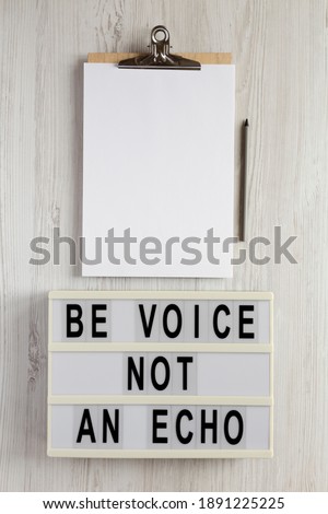 'Be voice not an echo' on a lightbox, clipboard with blank sheet of paper on a white wooden surface, top view. Flat lay, overhead, from above. 