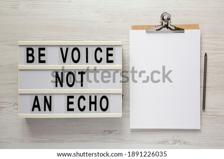 'Be voice not an echo' on a lightbox, clipboard with blank sheet of paper on a white wooden surface, top view. Flat lay, overhead, from above. 