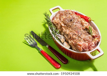 Raw pork roll stuffed with garlic and carrots in a ceramic roasting dish. Fresh meat, cutlery, spices, rosemary, garlic. Light green background, modern hard light, dark shadow, copy space