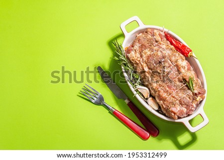 Raw pork roll stuffed with garlic and carrots in a ceramic roasting dish. Fresh meat, cutlery, spices, rosemary, garlic. Light green background, modern hard light, dark shadow, top view
