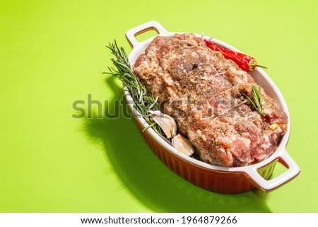 Raw pork roll stuffed with garlic and carrots in a ceramic roasting dish. Fresh meat, spices, rosemary, garlic. Light green background, modern hard light, dark shadow, copy space