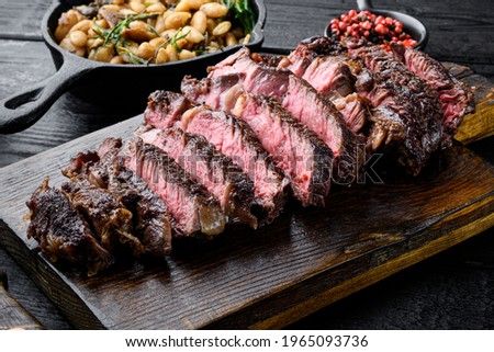 Sliced grilled meat steak Rib eye medium rare set, on wooden serving board, with white beans and rosemary in cast iron pan, on black wooden table background