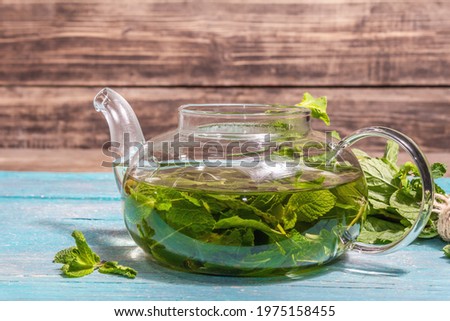 Mint tea from fresh leaves. Healthy hot drink in a glass teapot. Modern hard light, dark shadow, turquoise wooden boards background, place for text