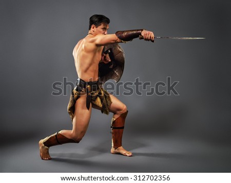 Half naked man in the image of ancient warrior strikes with melee weapons on a neutral grey background