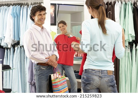 Woman trying on red top in clothes shop, looking at reflection in mirror, boyfriend looking on, smiling, rear view