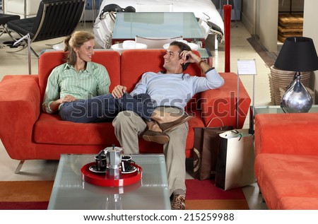 Couple testing new red sofa in furniture store, woman sitting with feet up on man\'s lap, smiling