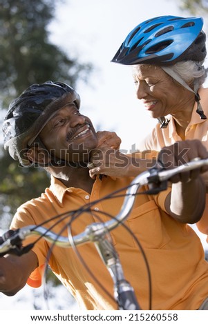 Active senior couple preparing to cycle in park, woman adjusting man\'s cycling helmet strap, smiling, close-up, low angle view (tilt)