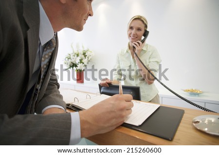 Businessman signing guestbook in reception area, female receptionist using telephone, smiling