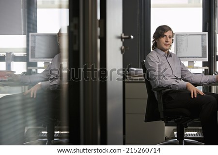 Businessman wearing telephone headset, sitting at desk in office, doorway view, side view, portrait
