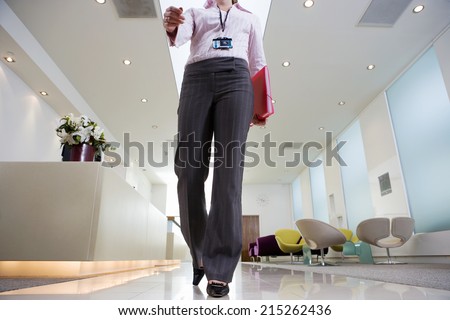 Businesswoman walking through lobby, carrying pink folder, low section, front view, surface level