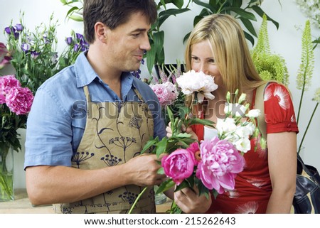Blonde woman smelling flowers in flower shop, male florist looking on, smiling, side view