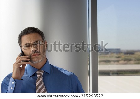 Mature businessman wearing spectacles, using mobile phone beside window, front view
