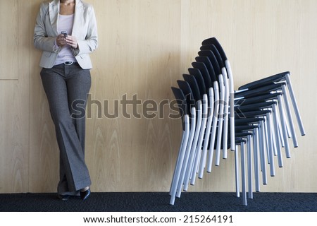 Businesswoman standing in conference room beside stack of chairs, holding mobile phone, low section