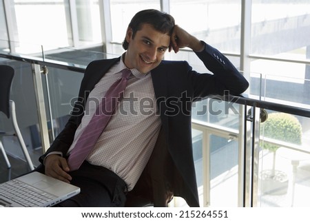 Businessman sitting in office reception area, laptop in lap, smiling, portrait