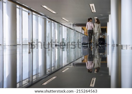 Two businesswomen walking with luggage in modern building, rear view, reflection on shiny floor
