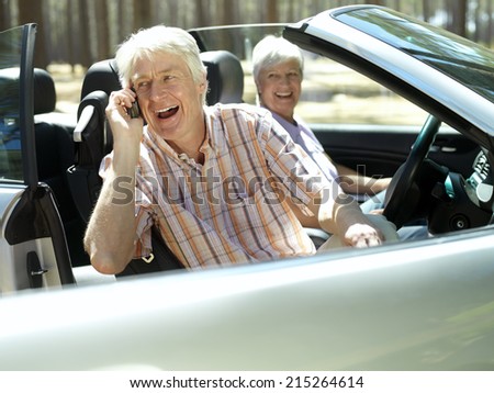 Senior couple sitting in convertible car, man using mobile phone, smiling, side view