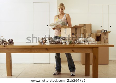 Woman moving house, packing crockery in cardboard boxes on dining room table, smiling, front view