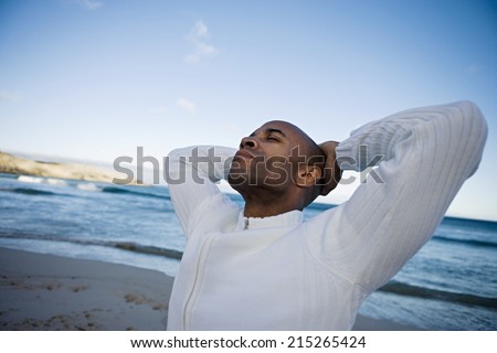 Man in white top standing on beach at sunset, hands behind head, looking up, thinking (tilt)