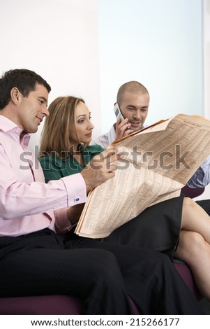 Three business colleagues sitting on sofa, man reading newspaper, second man using mobile phone