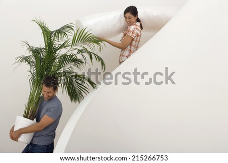 Couple moving house, man carrying large pot plant down staircase, woman carrying dust sheet, smiling