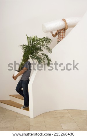 Couple moving house, man carrying large pot plant down staircase, woman carrying dust sheet