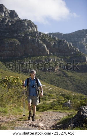 Mature man hiking on mountain trail, carrying rucksack, using hiking pole, looking at scenery, front view