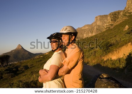Mature couple, in cycling helmets, standing on mountain trail, looking at scenery, man embracing woman, side view
