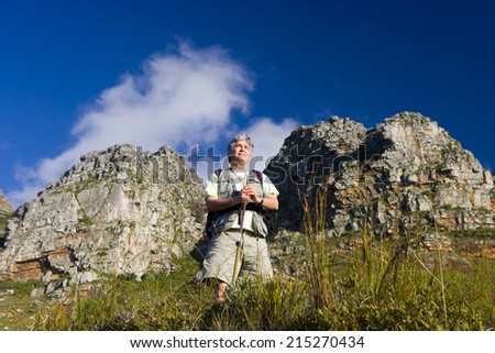 Mature man hiking on mountain trail, leaning on hiking pole, admiring scenery, smiling, low angle view