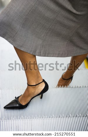 Businesswoman, in grey skirt and high heels, standing on escalator, close-up, low-section