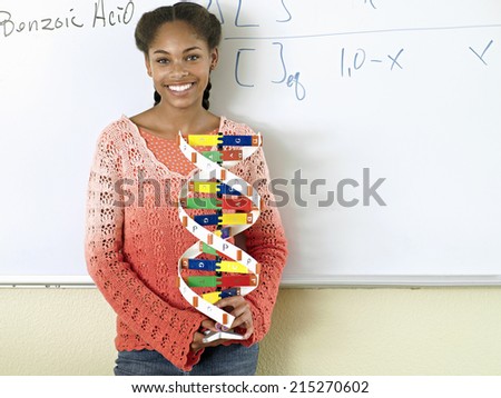 Teenage girl (15-17) standing beside whiteboard in classroom, holding DNA model, smiling, portrait