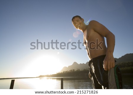 Man, in swimming shorts, standing on lake jetty at sunset, smiling, portrait, low angle view (lens flare, backlit)
