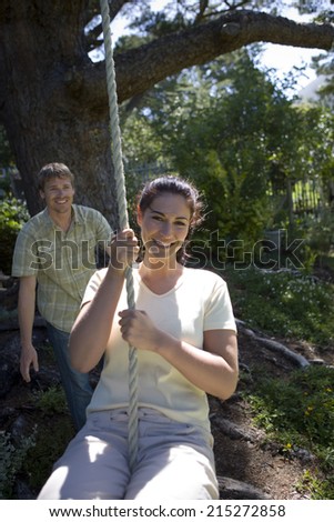 Woman swinging on garden rope swing, man pushing her in background, smiling, front view, portrait