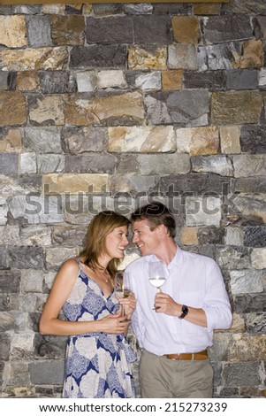 Couple flirting beside stone wall, holding glasses of white wine, smiling
