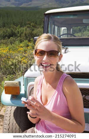 Young woman standing in front of parked jeep on dirt track, using mobile phone, smiling, portrait