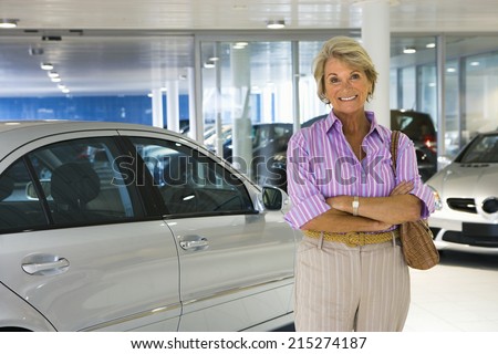 Senior woman standing beside new silver saloon car in large car showroom, smiling, portrait