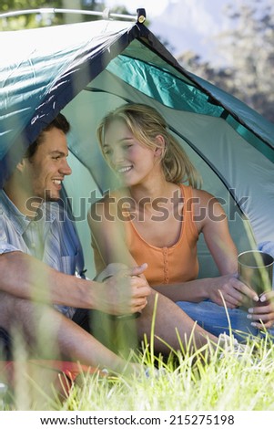 Young couple sitting in dome tent, holding camping mugs, smiling (differential focus)