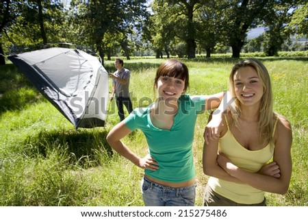 Young man assembling dome tent on camping trip in woodland clearing, focus on two female friends in foreground, smiling, portrait