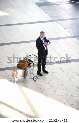 Senior businessman standing beside luggage trolley in airport, checking time on wristwatch, elevated view