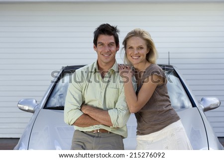 Couple leaning on bonnet of parked convertible car on driveway, smiling, front view, portrait