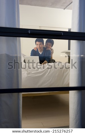 Young woman embracing young man using laptop on bed, smiling, view through window from outside