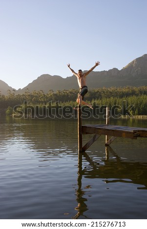 Man, in swimming shorts, jumping from jetty into lake, arms up, showing off, smiling