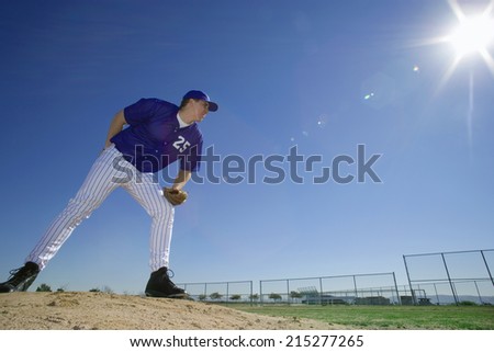 Baseball pitcher, in blue uniform, preparing to throw ball during competitive game, holding ball behind back, side view (lens flare, surface level)