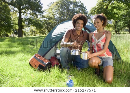 Young couple kneeling beside dome tent in woodland clearing, taking boiled kettle from camping stove, smiling, portrait