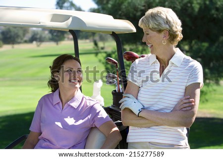 Two mature women talking on golf course, brunette sitting in golf buggy, blonde standing beside her, smiling