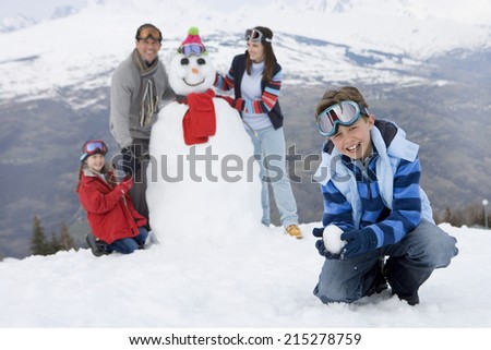 Boy crouching in snow, family by snowman in background, smiling, portrait
